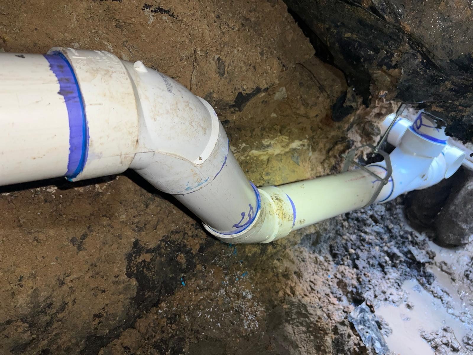 New PVC drain fittings hung in a hand-dug tunnel under a slab foundation.